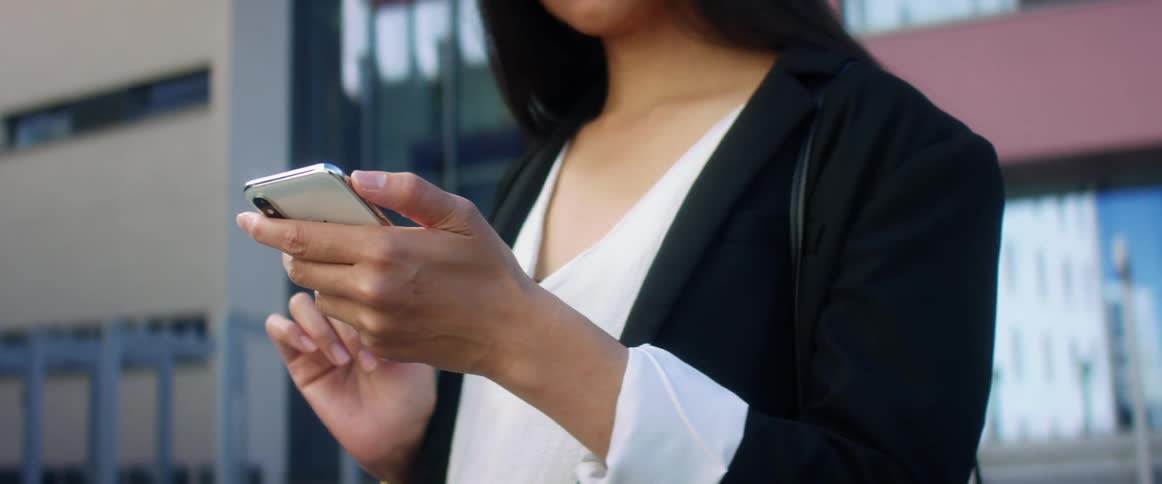 Asian woman with long hair a black blazer and a white shirt uses iphone xs in front of a building in the city street
