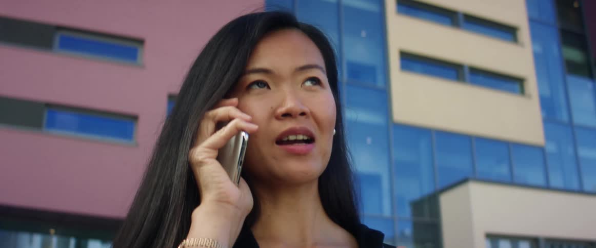 An asian woman talks on a smartphone while standing outside of a large building in a city during the day