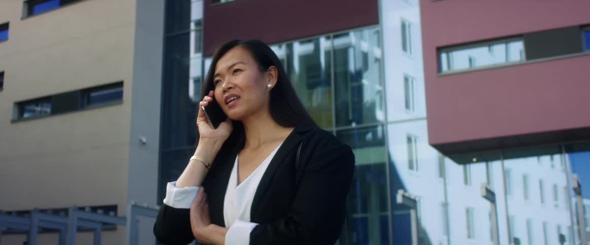 Asian woman with with long hair and a black blazer talks on the smartphone in front of a high rise buildings