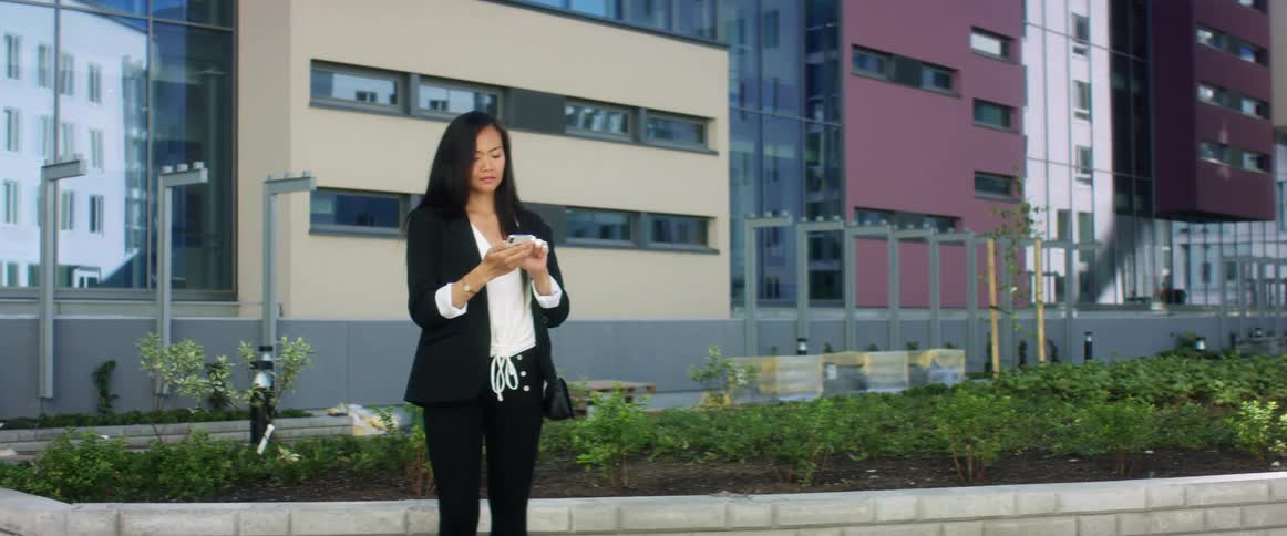 An asian woman dials on her smartphone before lifting the phone to her ear while standing outdoors in a metropolis during the day
