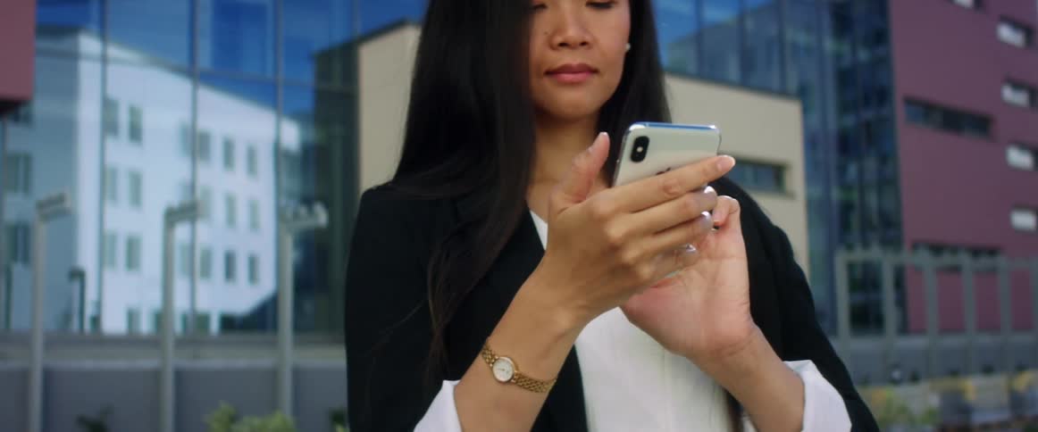 Asian woman with long hair and a black blazer uses an iphone smartphone in the city street