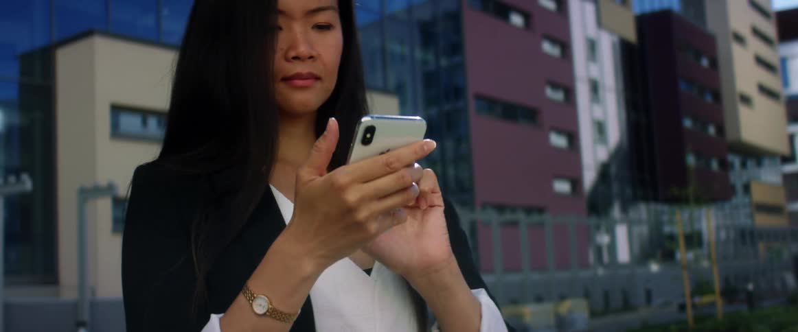 Asian woman with long straight hair and a black blazer uses iphone smartphone in the street with buildings in the background