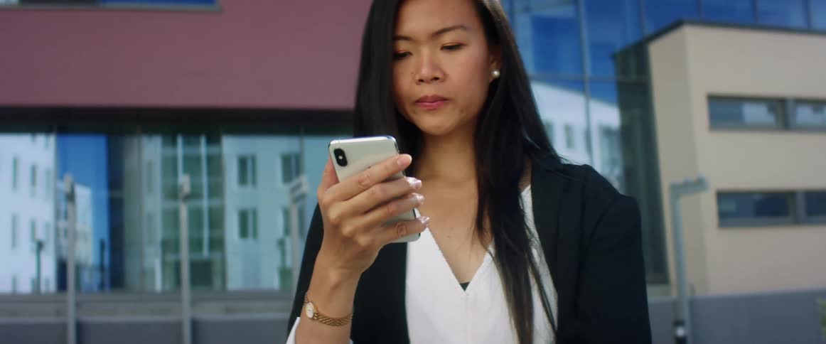 A young woman standing outside office buildings in a downtown area looks at her cell phone and scrolls through it