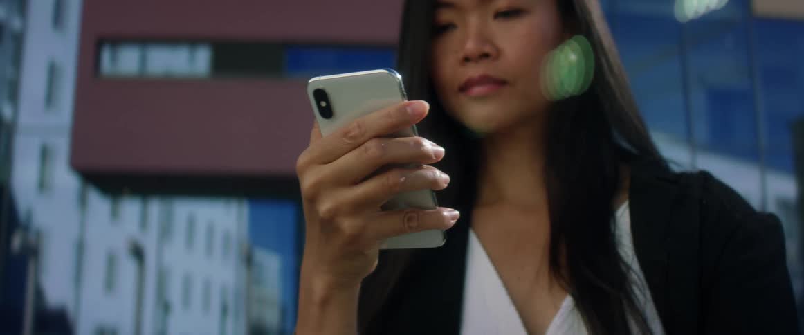 Asian woman with long straight hair a black blazer and a white shirt uses smartphone in the city street with high rise buildings in the background