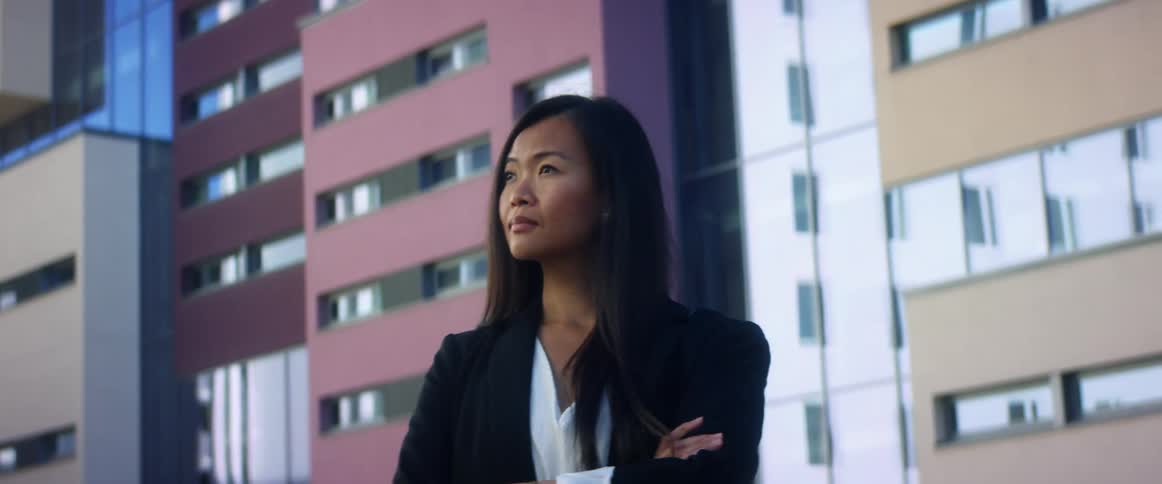 Asian woman with long hair and blazer jacket stands arms crossed in front of high rise buildings