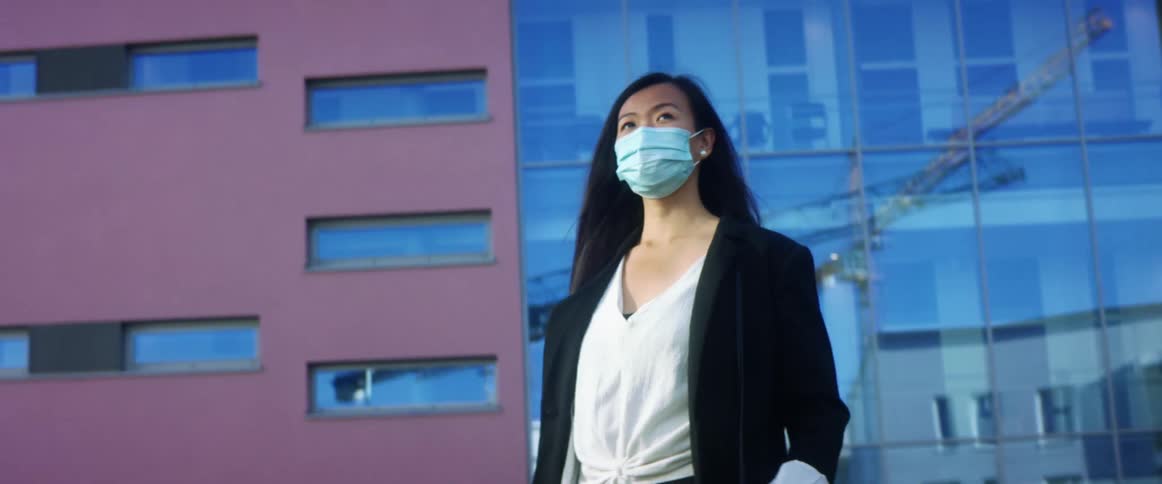 Woman wearing blazer and face mask walks away from building with reflective glass