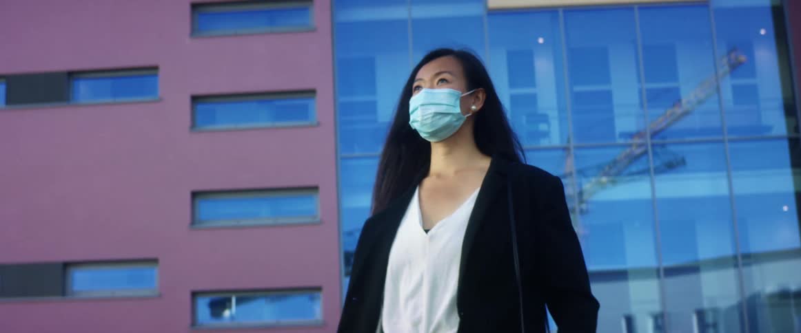 An asian woman in formal clothing and a face mask walks around from a tall building in a city on a sunny day