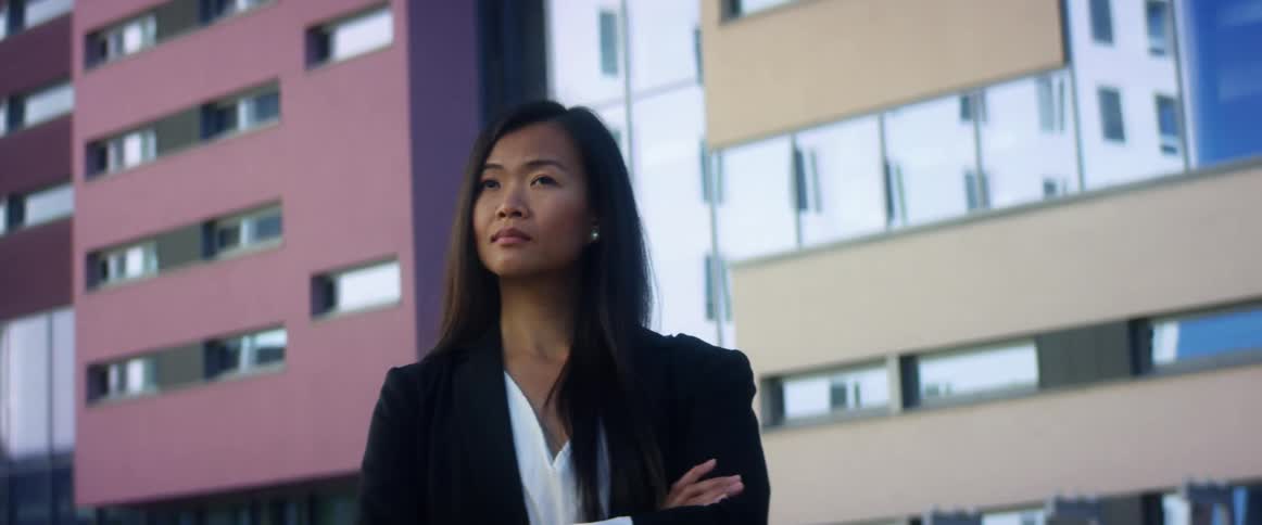 Asian woman with long hair and a blue mask stands arms crossed and looks around in front of tall buildings
