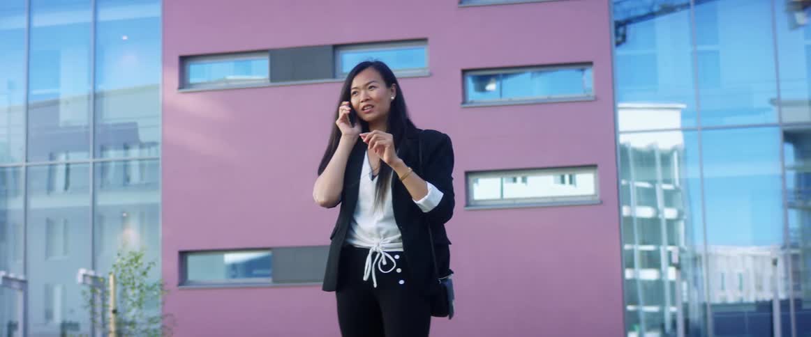Asian woman with long hair a black blazer and a white shirt talks on the smartphone in front of a building