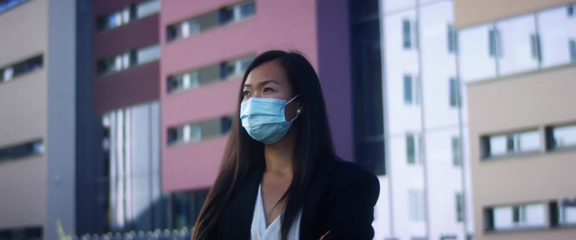 Asian woman with long hair and a blue mask stands arms crossed in front of tall buildings