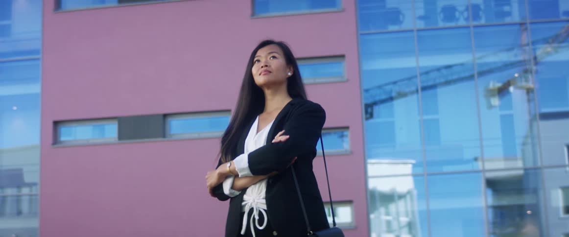 Woman walks next to the city buildings with her arms crossed and a purse hanging over her shoulder