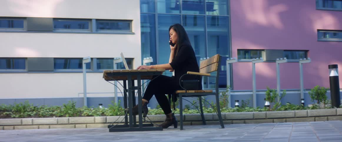 An asian woman sitting at a table outdoors talks on her smartphone while typing on a laptop with her free hand