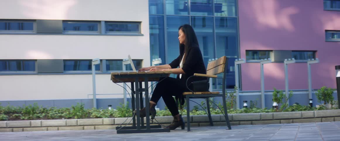 Asian woman with a white shirt and a black jacket sits at a wood table and opens a laptop to use outdoors