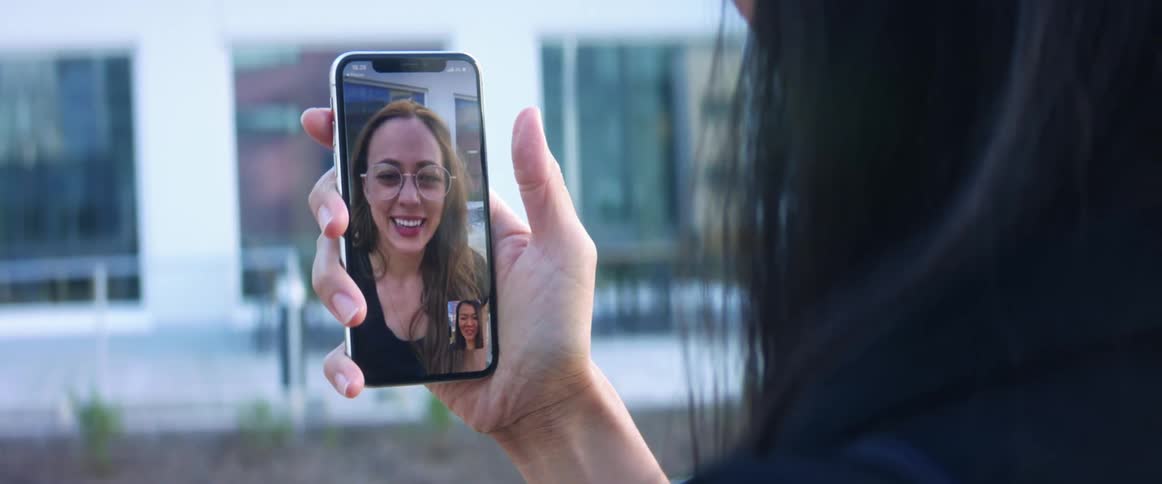A woman standing outdoors in a city during the day video chats with another woman