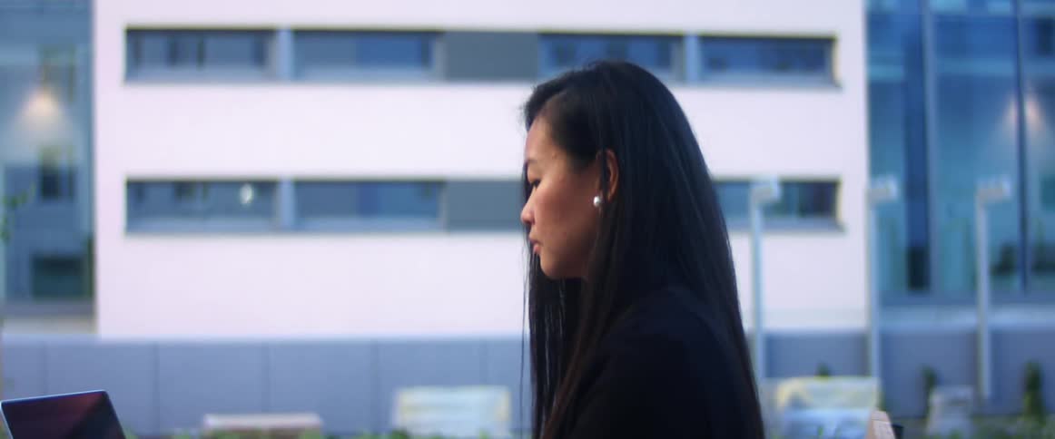 A woman sitting outside an office building in a downtown area types on her laptop