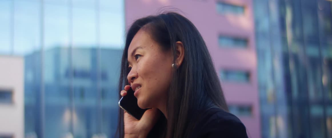 Asian woman with long hair talks on the smartphone in city street with tall buildings in the background