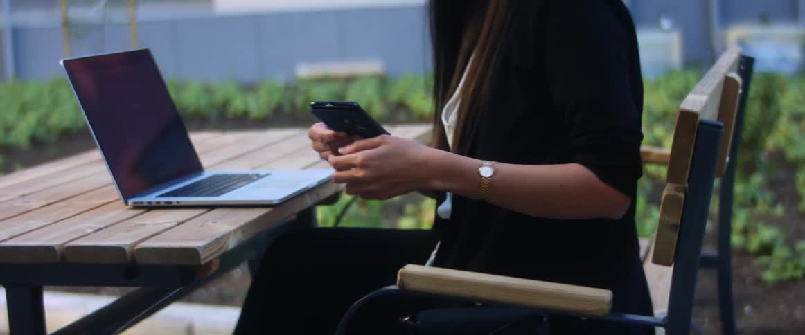 A woman sitting outside with her laptop takes her phone out of her purse and scrolls through it