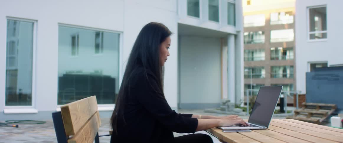 A woman sits at a table outside a large building with a laptop