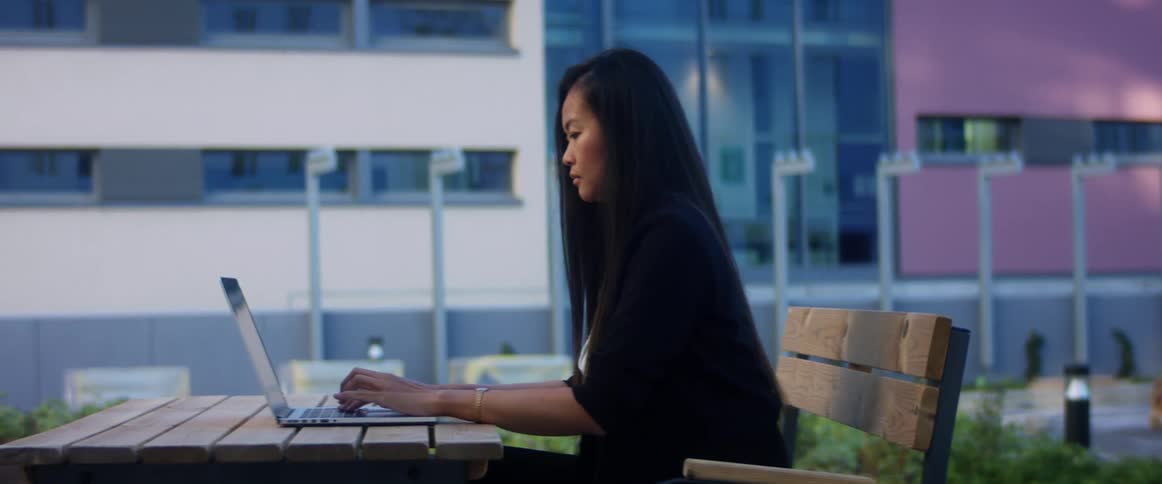 A woman sits at a wooden patio table and types on her laptop computer
