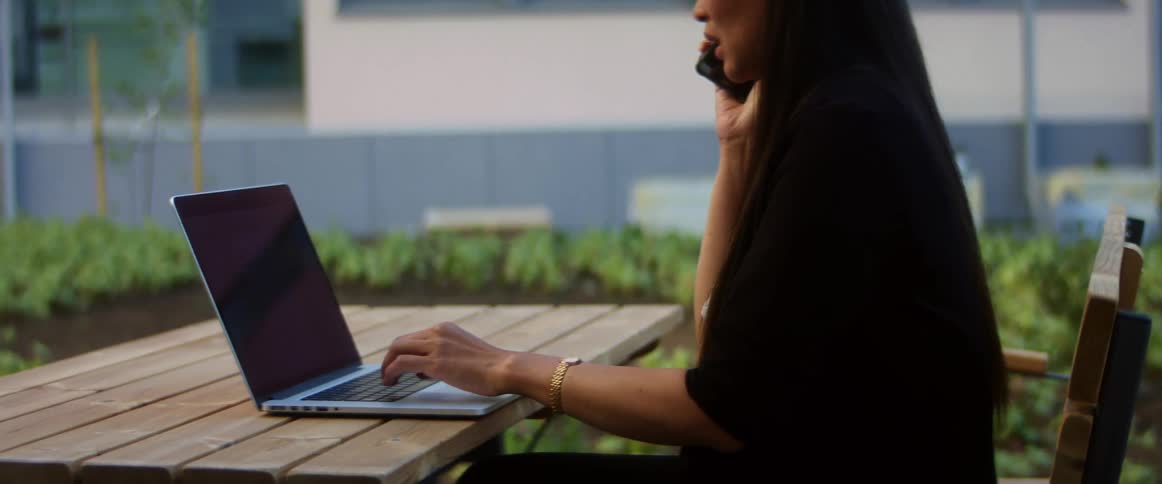 A woman sits at a wooden table outdoors typing on a laptop with one hand while holding a smartphone to her face with the other