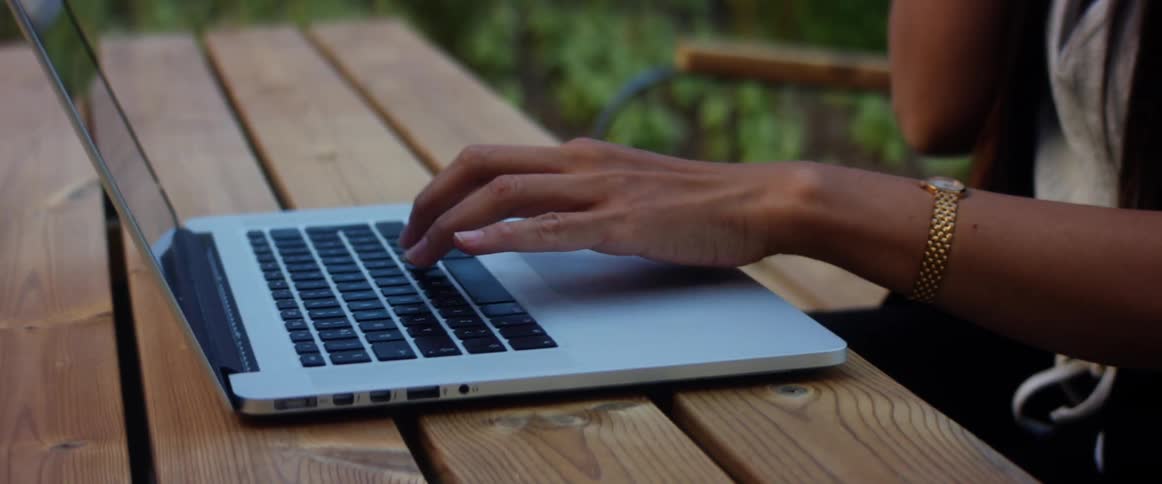 Woman sits outdoors at wooden picnic table typing on laptop