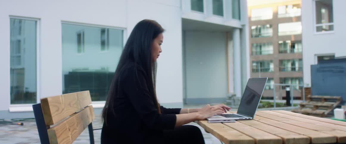 A woman sits at a table and bench with a laptop outside of a large building