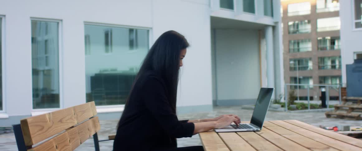 A woman sitting on a bench next to a table typing on a laptop