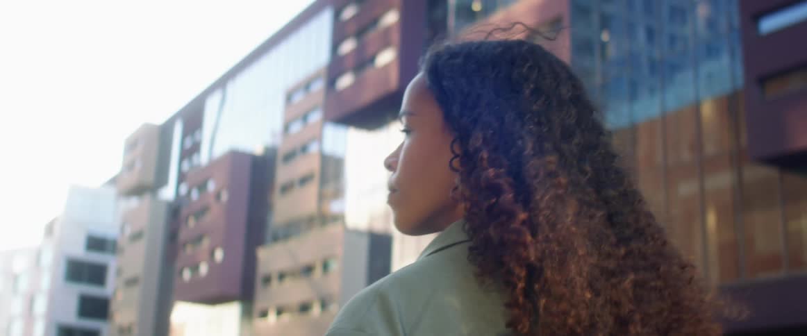 Black woman with curly hair and a blue overcoat walks down the street with buildings in the background