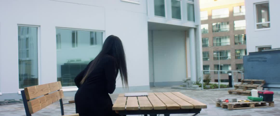A young woman sits down at a table in an apartment courtyard and opens up her laptop