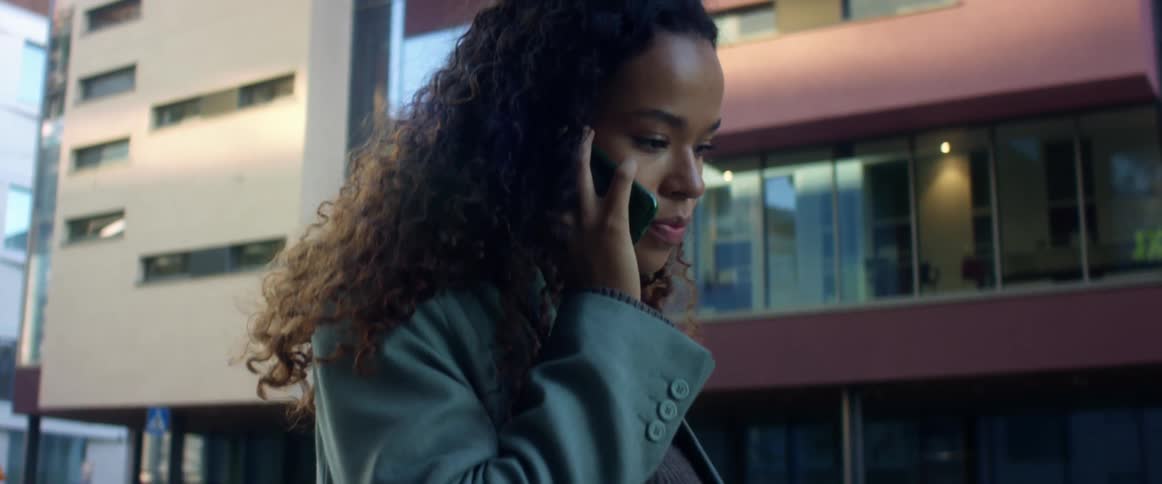 Black woman with curly hair and a blue top coat talks on the smartphone while walking down the city street