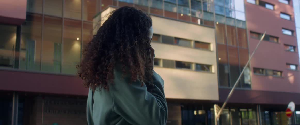 Black woman with curly hair and a blue topcoat talks on the smartphone while walking down the city street