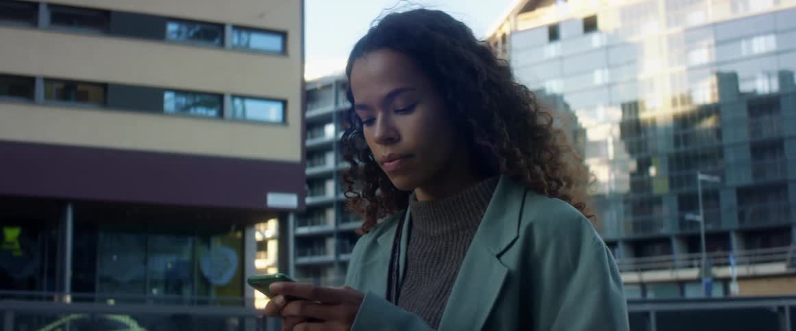 A young black woman standing outdoors in a metropolis during the day looks up at the buildings around here before turning her focus to the smartphone in her hands