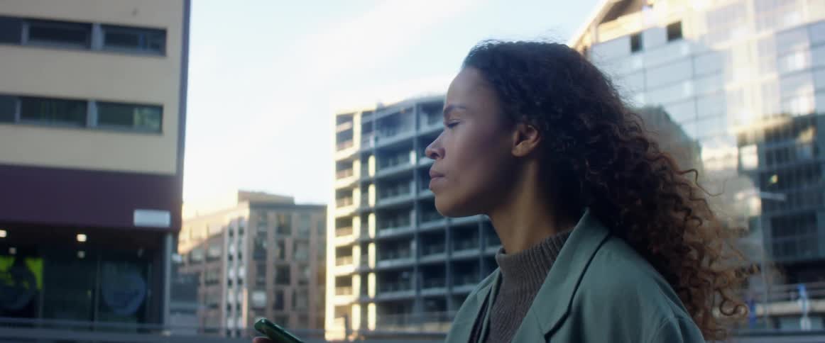 Black woman with curly hair and a blue coat walks down the street and uses a smartphone