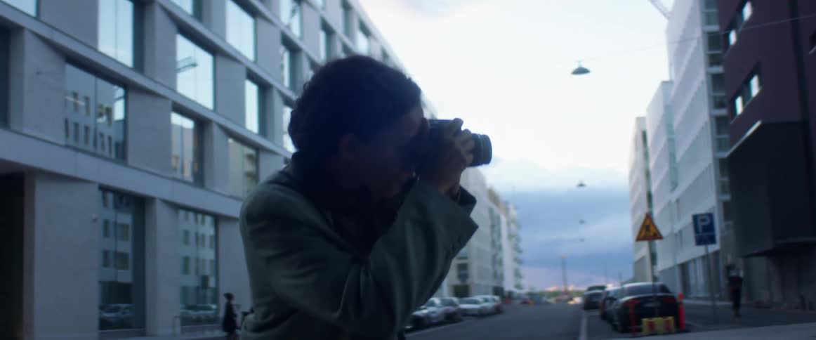 Black woman with curly hair and a blue top coat takes photographs with a camera and checks them in the street