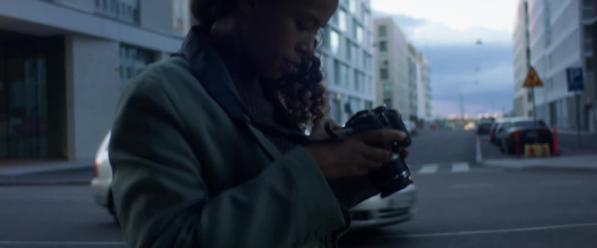Black woman with curly hair and a blue top coat takes photographs and checks them in the street with buildings in the background at dusk