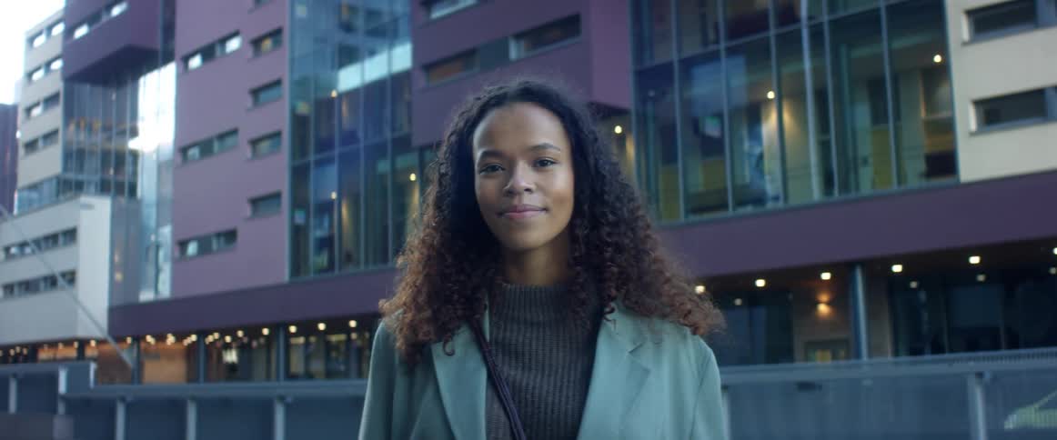 Black woman with curly hair and a blue coat smiles warmly and faces the camera with eye contact in the street with buildings in the background