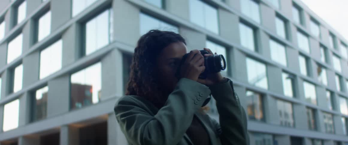A woman stands outside of a city building and puts a camera to her face