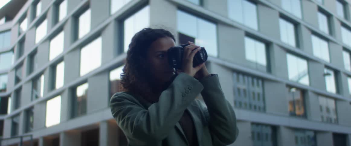 Black woman with curly hair and a blue top coat takes photographs in the street with buildings in the background