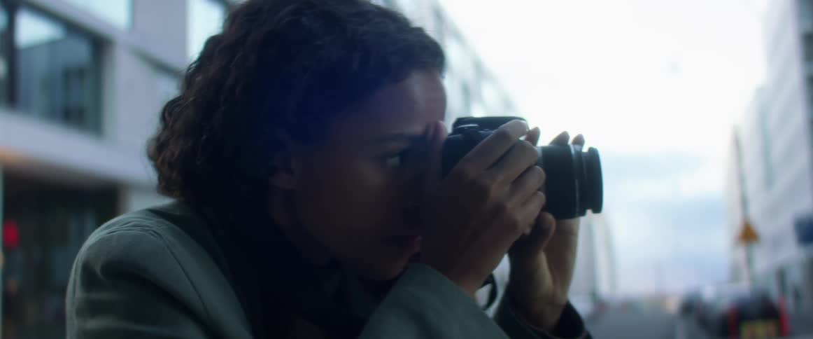 Black woman with curly hair and a light blue overcoat takes photographs with a camera in the city street at dusk