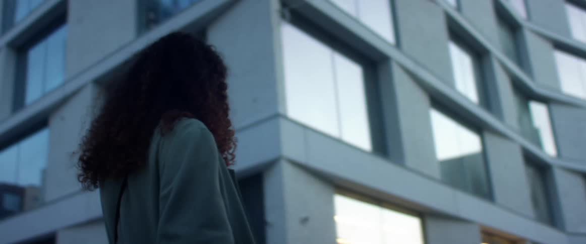 Black woman with curly hair and a blue topcoat talks on the smartphone while walking down the city street
