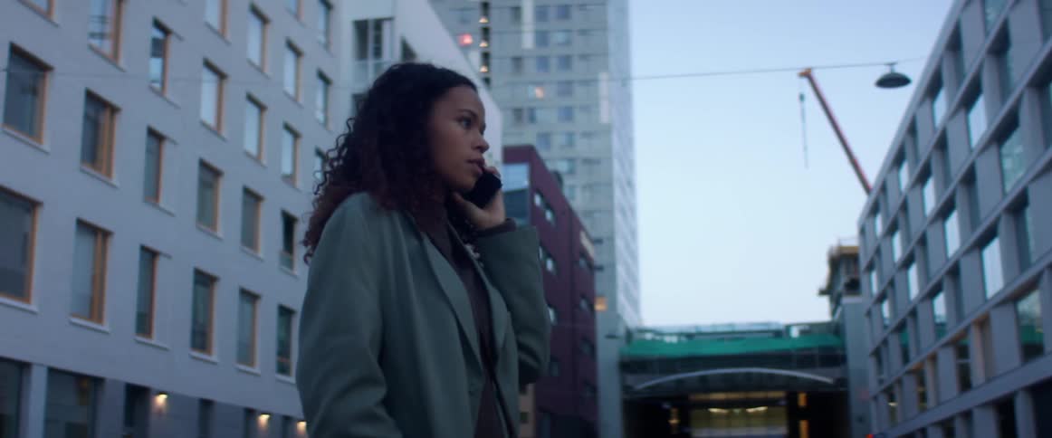 Black woman with curly hair and a blue topcoat talks on the smartphone while walking down the city street