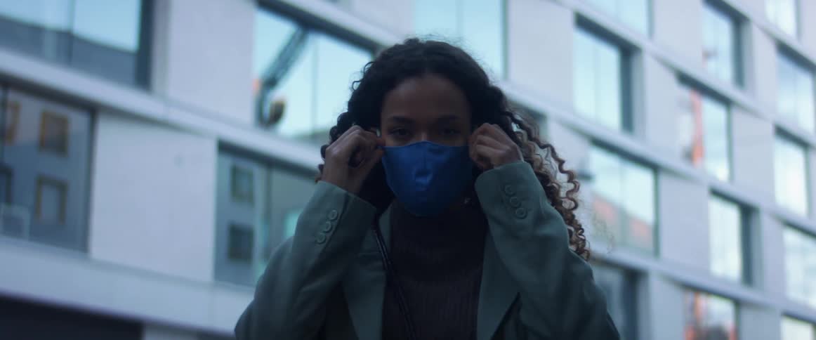 Black woman with curly hair and a blue coat wears a mask in front a building in the city street
