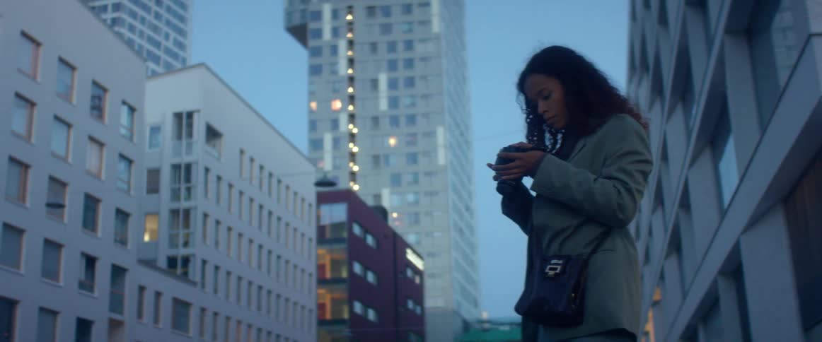 Black woman with curly hair and a blue top coat takes photographs and checks them in the street with high rise buildings in the background at dusk