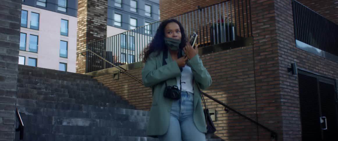 Black woman with curly hair a light blue coat jeans and a pair of white shoes goes down the street stairs and wears a grey mask