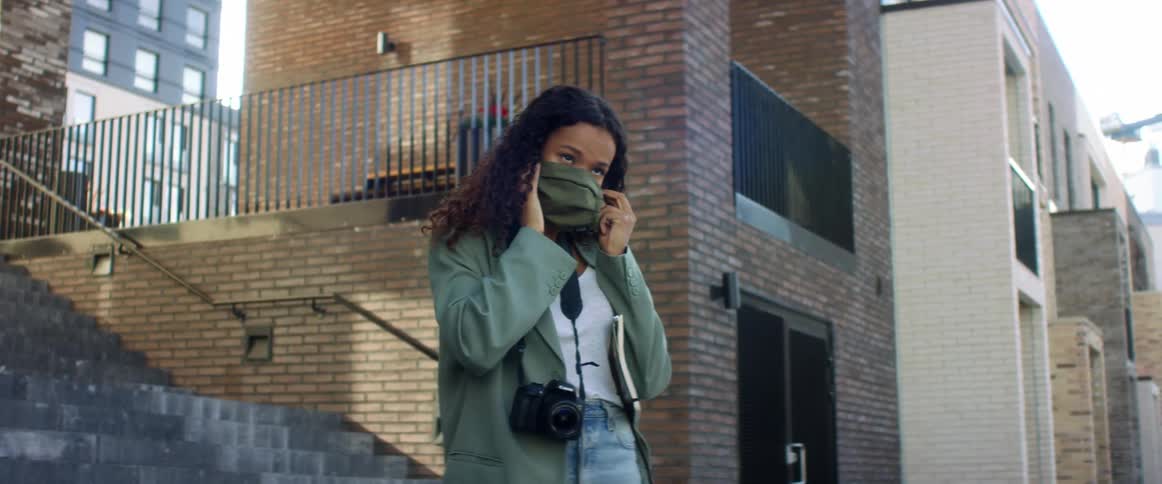 Black woman with curly hair jeans and a blue coat wears a grey mask in the city street