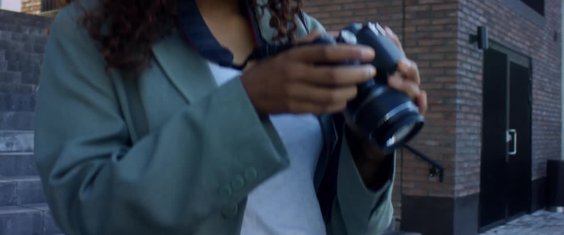 A young black female photographer wearing a medical mask examines her camera as she stands outdoors in a city during the day