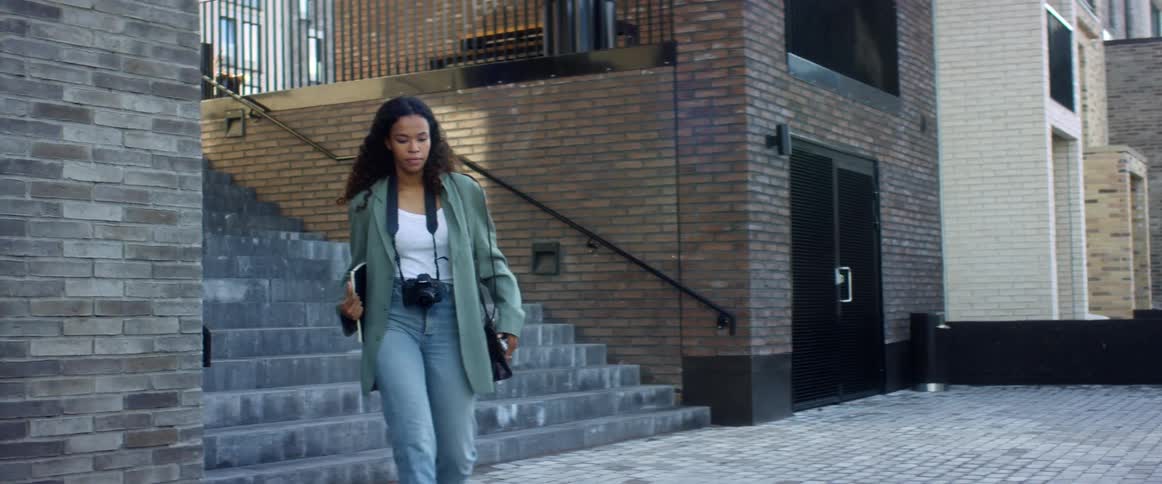 A teenage girl carrying a tablet and with a camera around her neck walks down some stairs in a city
