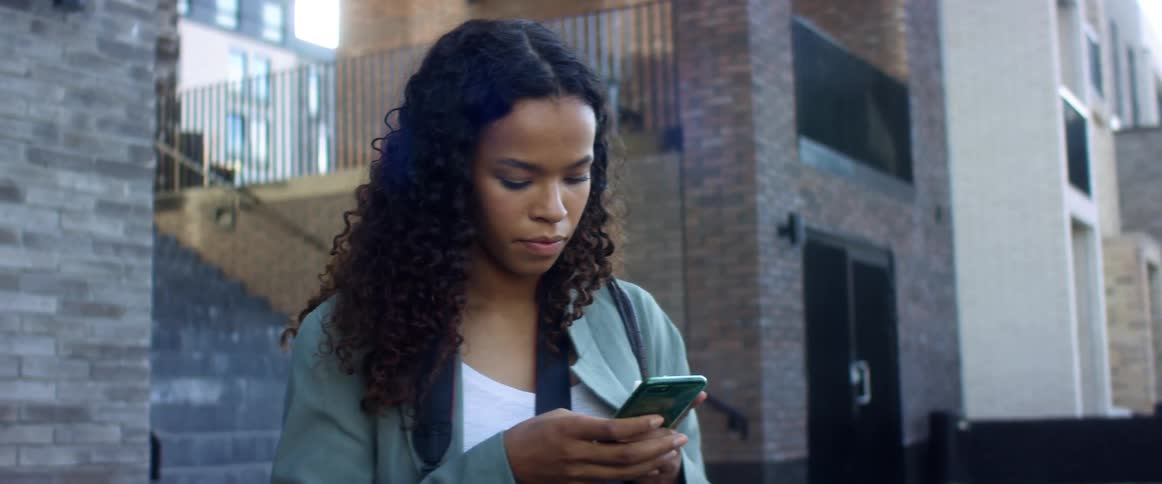 A woman stands outside of a city building and pulls her cell phone out of her jacket pocket