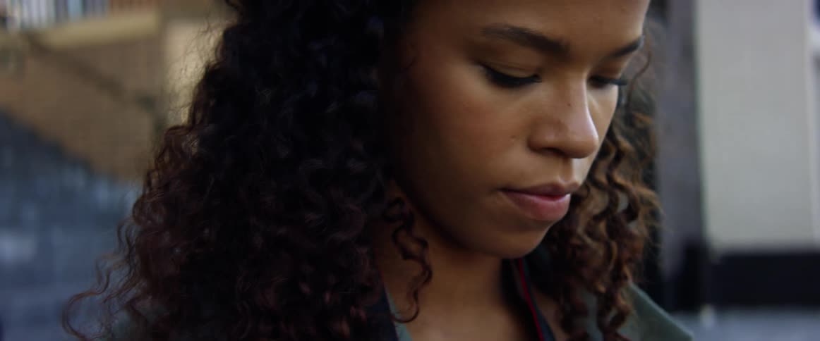 A young black woman looks downward in contemplation as she stands outdoors in a metropolis during the day
