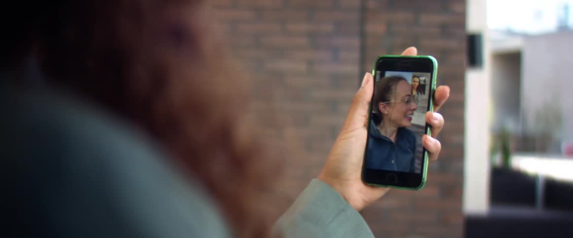 A pair of women use their smartphones to video chat with one another while standing outdoors during the day
